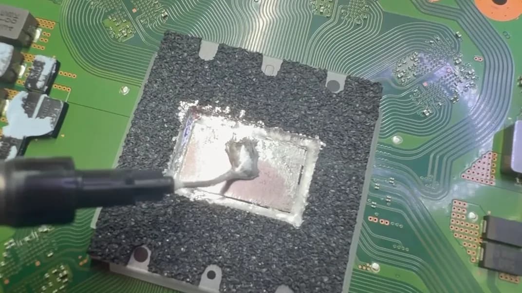 Nahaufnahme des PS5-Prozessors unter dem Mikroskop: frisches Flüssigmetall wird gleichmäßig auf dem Die verteilt, ringsum der schwarze Schaumstoffrahmen, der das Metall an Ort und Stelle hält