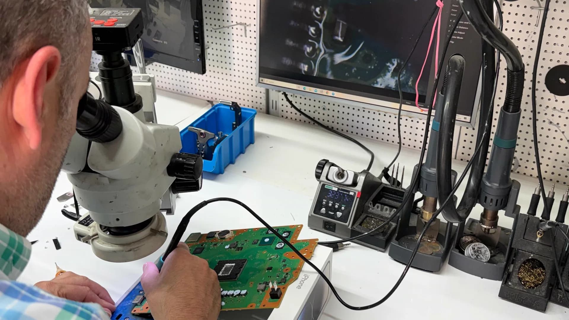 Techniker am Stereo-Mikroskop in der Wiesbadener Werkstatt: vor ihm ein ausgebautes PS5-Mainboard, daneben Lötstation und Heißluft, im Hintergrund der Monitor mit dem Mikroskopbild